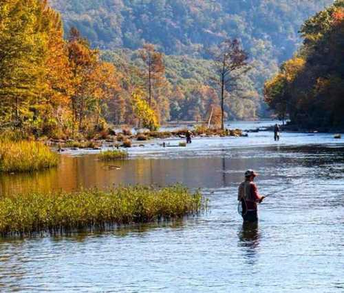 A serene river scene with fishermen wading through water, surrounded by autumn foliage and mountains in the background.