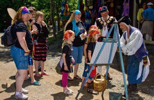 A group of people in colorful attire watches a man painting on an easel at a lively outdoor event.