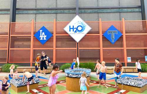 Children playing in a splash pad area at a water park, with a sign for H2O and logos for LA and another team in the background.