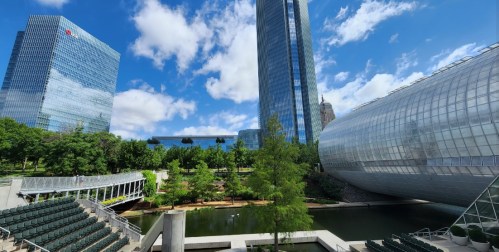 A modern urban landscape featuring glass buildings, greenery, and a reflective pond under a blue sky with clouds.
