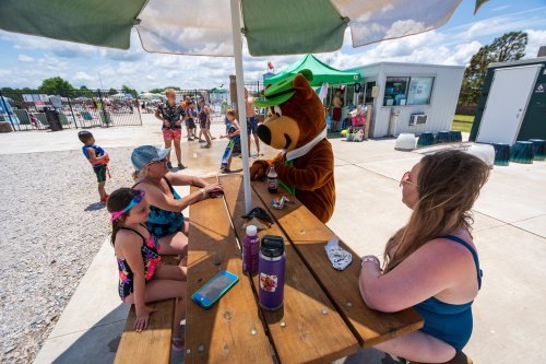 A costumed bear interacts with children and adults at a picnic table near a pool on a sunny day.