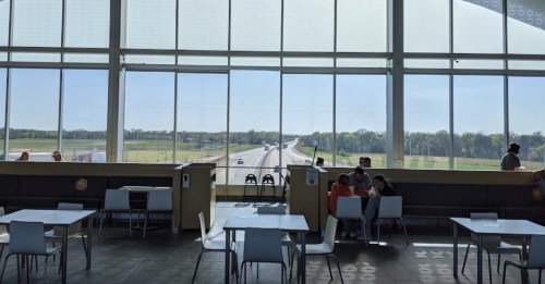 Bright interior of a café with large windows overlooking a highway and green landscape. Tables and chairs are visible.