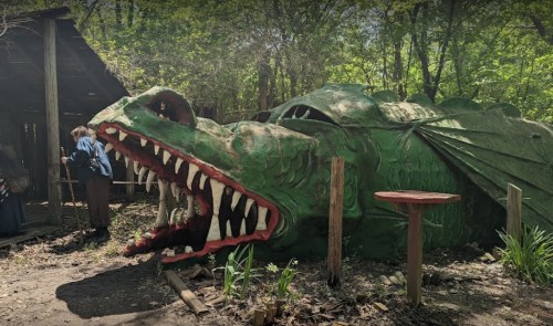 A large, green dragon sculpture lies on the ground in a wooded area, with people nearby exploring the site.