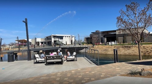 A group of people sits by a water feature in a park, with shops and a clear blue sky in the background.
