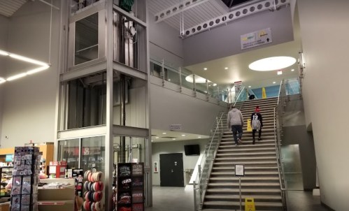 Interior view of a modern building featuring stairs, an elevator, and people walking upstairs. Bright and spacious design.