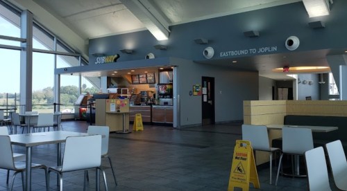 Interior of a Subway restaurant with seating, a counter, and a sign for "Eastbound to Joplin." Caution sign visible.