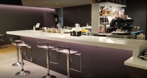 A modern bar with a sleek white countertop, high stools, and a well-stocked back shelf of drinks and snacks.