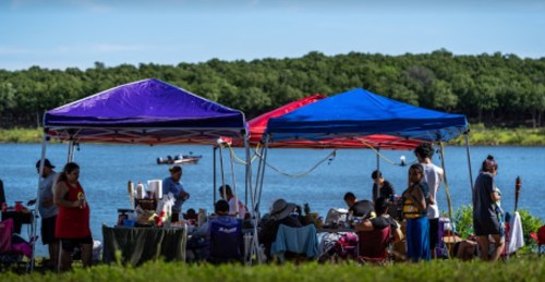 People gather under colorful tents by a lake, enjoying food and activities on a sunny day.