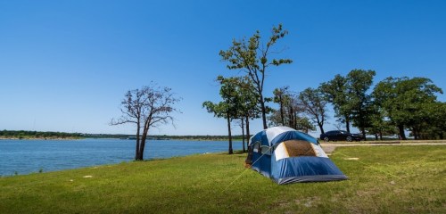 A blue and tan tent set up on green grass by a calm lake, with trees and a clear blue sky in the background.