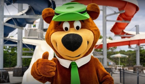 A friendly bear mascot in a green hat and tie gives a thumbs up at a water park with slides in the background.