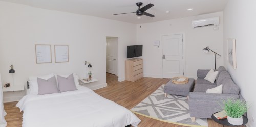 A bright, modern room featuring a bed, sofa, TV, and minimalist decor with wooden flooring.