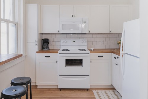 A modern kitchen with white cabinets, a stove, microwave, and a coffee maker, featuring wooden countertops and two stools.