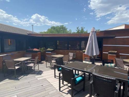 A spacious outdoor patio with tables, chairs, and an umbrella, surrounded by greenery and a clear blue sky.