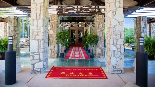 Entrance of a building with stone pillars, glass doors, and a red welcome mat surrounded by greenery.