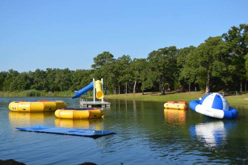 Colorful inflatable water toys and a slide on a calm lake, surrounded by green trees under a clear blue sky.