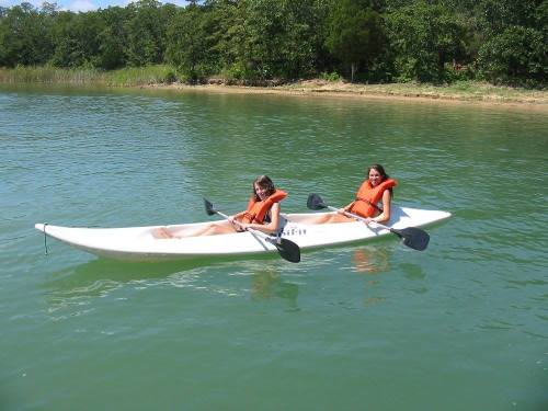Two people in orange life jackets paddle a kayak on a calm green lake surrounded by trees.