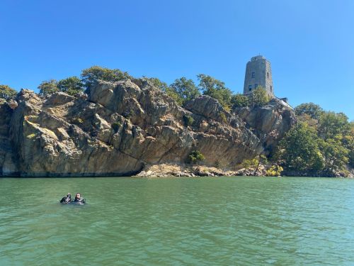 A rocky shoreline with a tower in the background, while two divers float in the green water below a clear blue sky.
