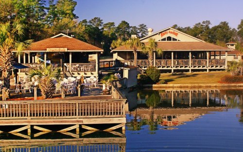 A scenic view of a waterfront restaurant with outdoor seating, surrounded by palm trees and calm water.
