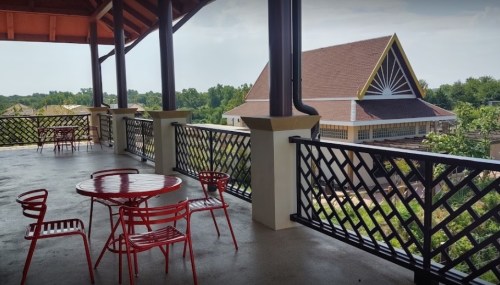 A spacious balcony with red chairs and tables overlooking a green landscape and a building with a distinctive roof.