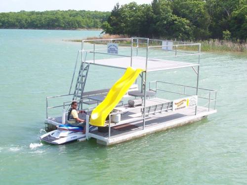 A floating platform on a lake with a slide, a ladder, and a person on a jet ski nearby. Green trees in the background.