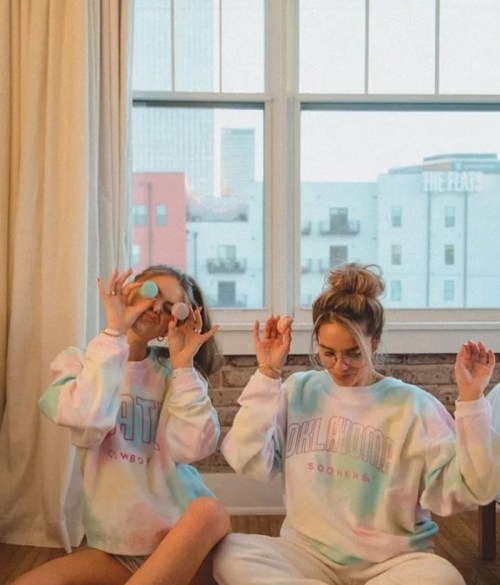 Two women in colorful sweatshirts playfully pose with sunglasses indoors, sitting by a window with a city view.