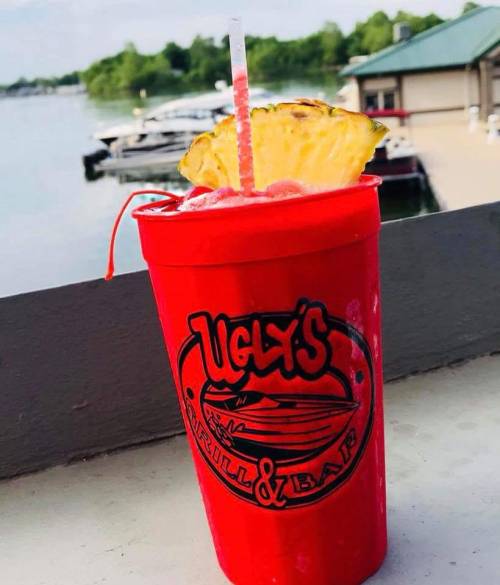 A vibrant red cup from Ugly's Grill & Bar filled with a colorful drink, garnished with a pineapple slice, by the water.