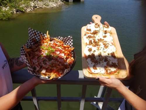 Two people hold plates of loaded fries and nachos against a scenic outdoor backdrop by the water.