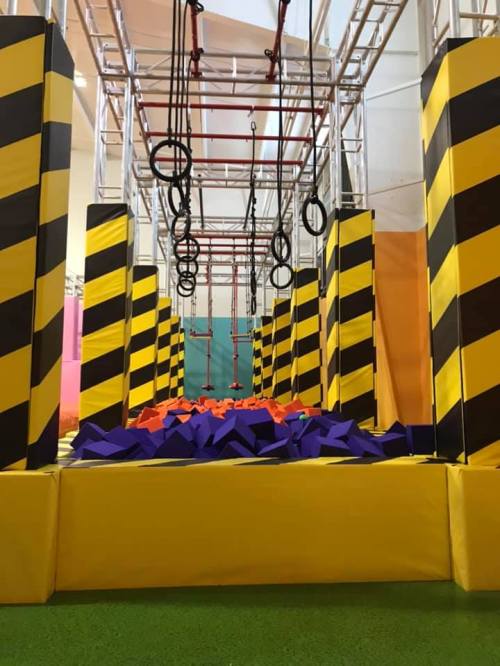 An indoor obstacle course with yellow and black striped pillars, rings, and a foam pit filled with purple and orange blocks.