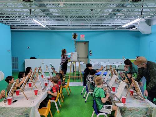 A colorful art studio with children painting on easels, guided by an instructor, surrounded by bright blue walls.
