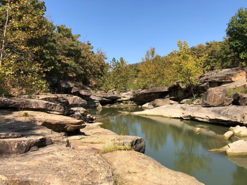 A serene river flows between rocky banks, surrounded by trees under a clear blue sky.