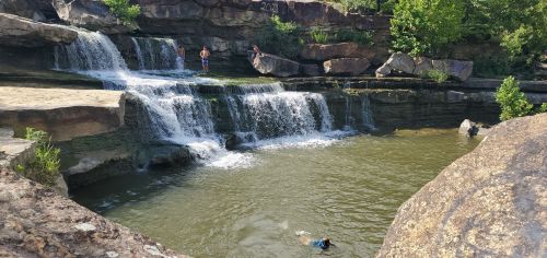 A serene waterfall cascades into a calm pool, with people enjoying the water and lush greenery surrounding the scene.