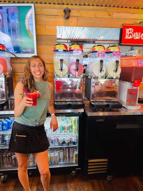 A smiling woman holds a drink in front of colorful slushie machines in a casual setting.