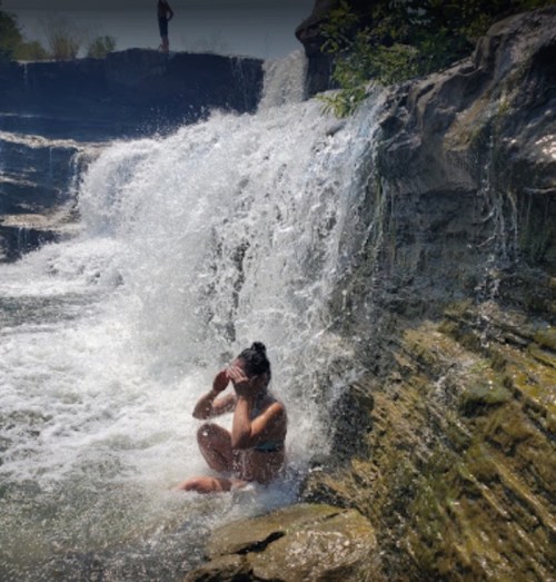 A person sits by a waterfall, splashing water around, surrounded by rocky terrain and lush greenery.