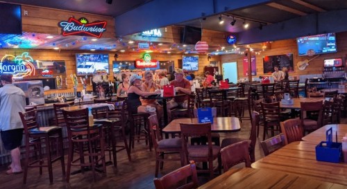 A lively restaurant interior with wooden tables, bar stools, and neon signs, featuring patrons enjoying food and drinks.