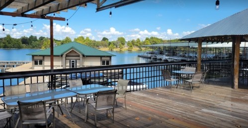 A scenic view of a lakeside deck with tables, overlooking a marina and lush greenery under a blue sky.