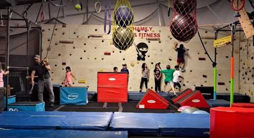 Children and adults engage in various activities at a gym with climbing walls and obstacle courses.