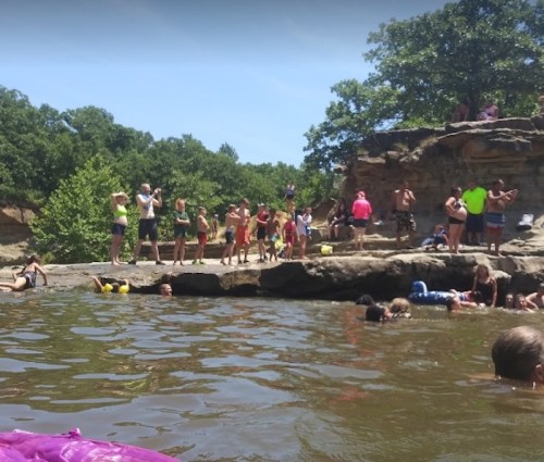 A lively scene at a swimming area with people enjoying the water and sun on rocks surrounded by trees.