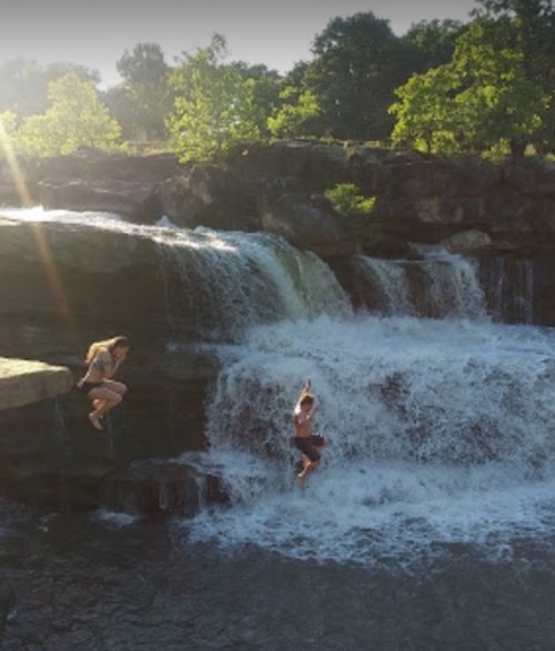 Two people jumping off rocks into a waterfall, surrounded by lush greenery and sunlight.