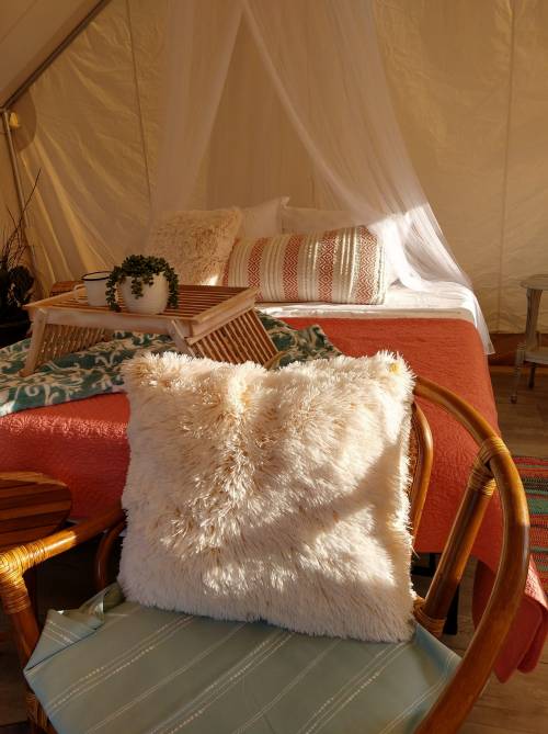 Cozy glamping setup featuring a bed with decorative pillows, a plush throw, and a chair with a fluffy cushion.