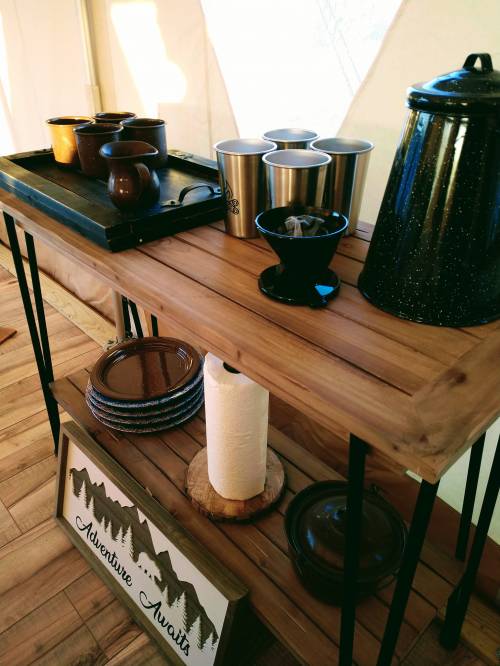A wooden table with coffee mugs, a kettle, and plates, featuring a decorative sign that reads "Adventure Awaits."