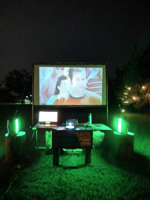 Outdoor movie setup at night with a projector screen, laptop, and green lights illuminating the area.