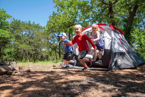 Three children run out of a tent in a sunny campsite, surrounded by trees and a rocky ground.