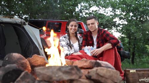 A couple sits by a campfire, wrapped in a blanket, with a tent and trees in the background.