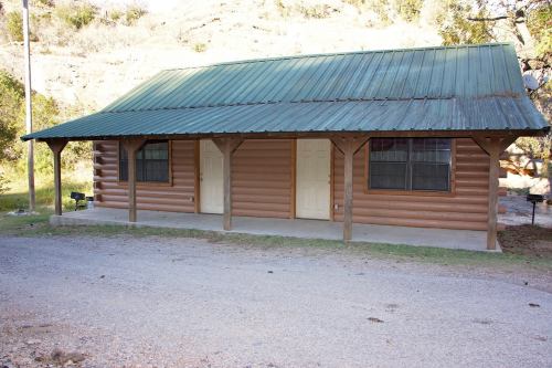A rustic log cabin with a green metal roof, featuring two doors and a porch, set in a natural landscape.