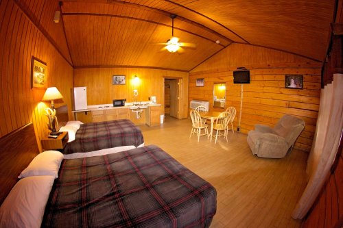 Cozy wooden cabin interior with two beds, dining area, kitchenette, and a comfortable chair. Warm lighting enhances the ambiance.