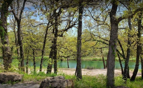 A serene river scene framed by trees, with a sandy area and a picnic table visible along the water's edge.