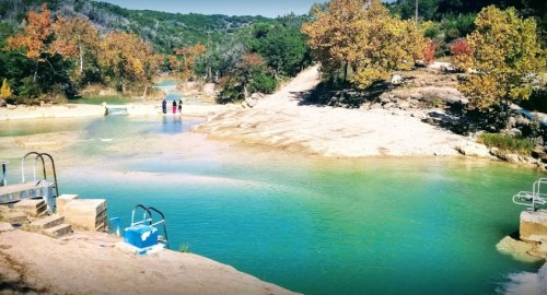 A serene river scene with clear turquoise water, surrounded by trees and people enjoying the outdoors.