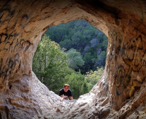 A person climbs through a rocky opening, surrounded by trees and graffiti on the stone walls.