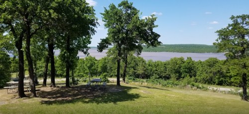A scenic view of a river surrounded by green trees and picnic tables under a clear blue sky.