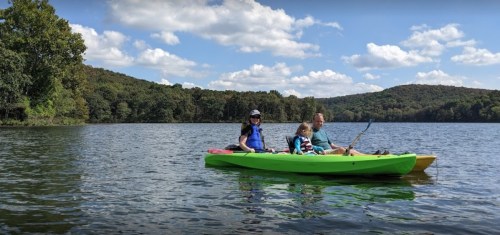 A family kayaking on a calm lake surrounded by trees and blue skies with fluffy clouds.